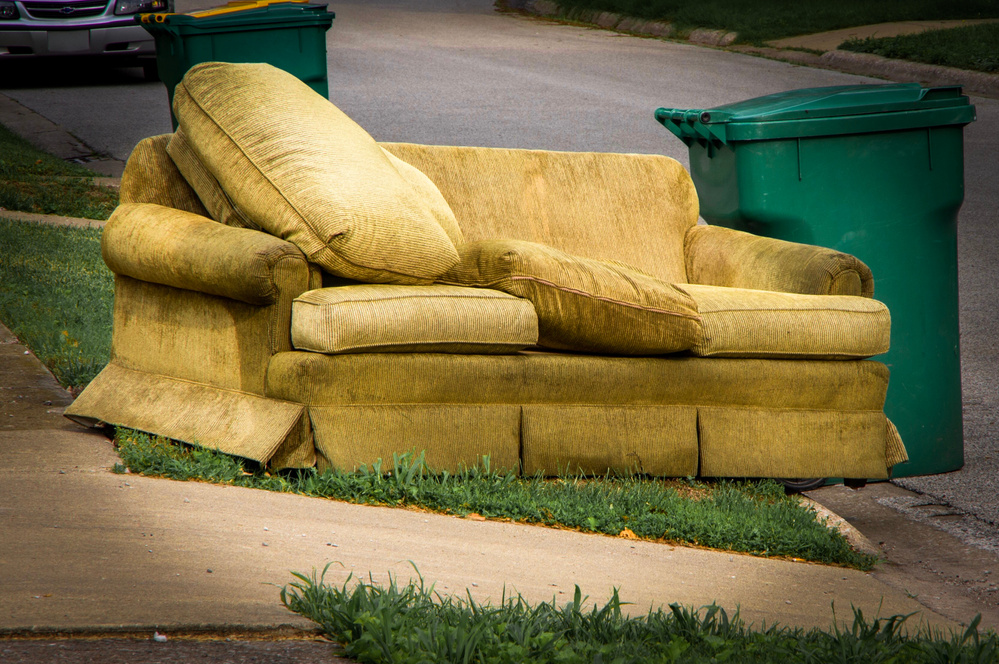 Old couch sitting along curb beside garbage container on trash day